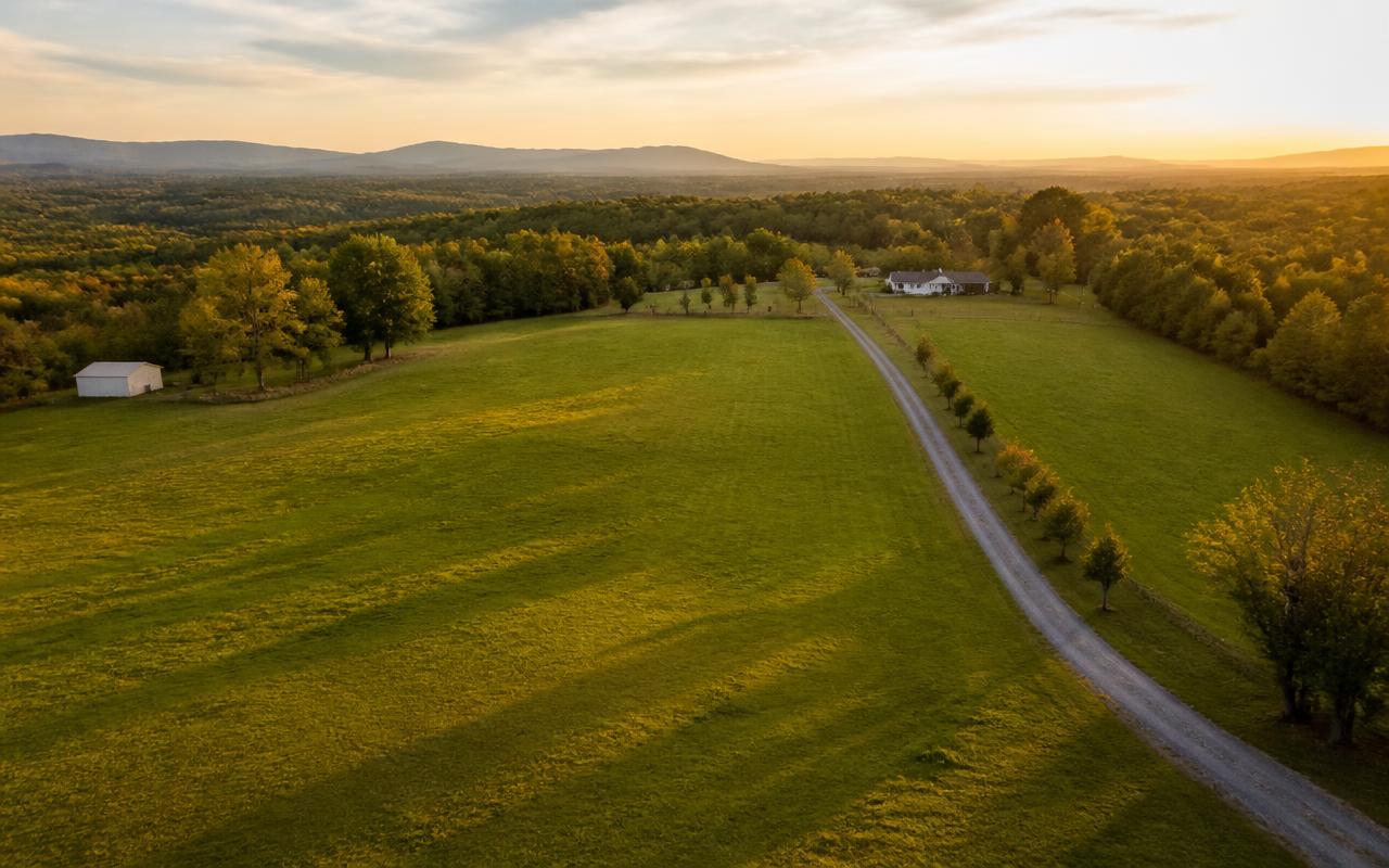 Aerial view of open rural acreage at golden hour with a gravel driveway and tree line